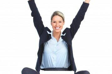 Extraordinary Woman sitting on the ground in front of her laptop with her arms up in celebration while she smiles at the camera