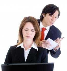 micromanager1 woman working on her computer with a man looking over her shoulder writing something down on a notepad