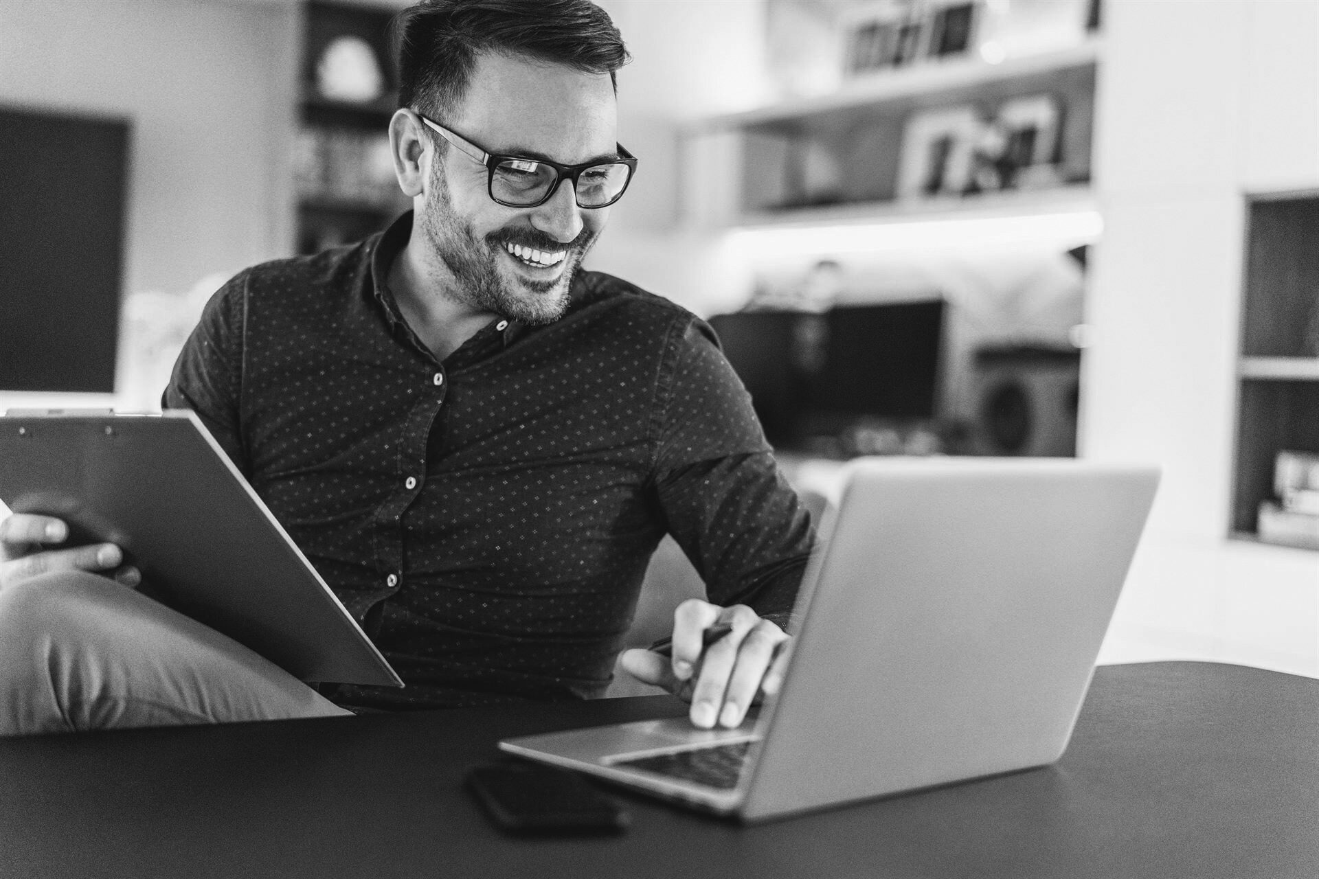 iStock-1213316704 Man sitting at a desk staring at his computer smiling, he is holding a clipboard in one hand and a pen in his other.