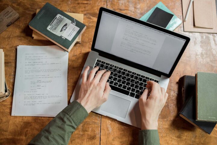 How to hire a copywriter birdseye view of a laptop, book, and sheet of paper. with hands typing on keyboard How to hire a copywriter